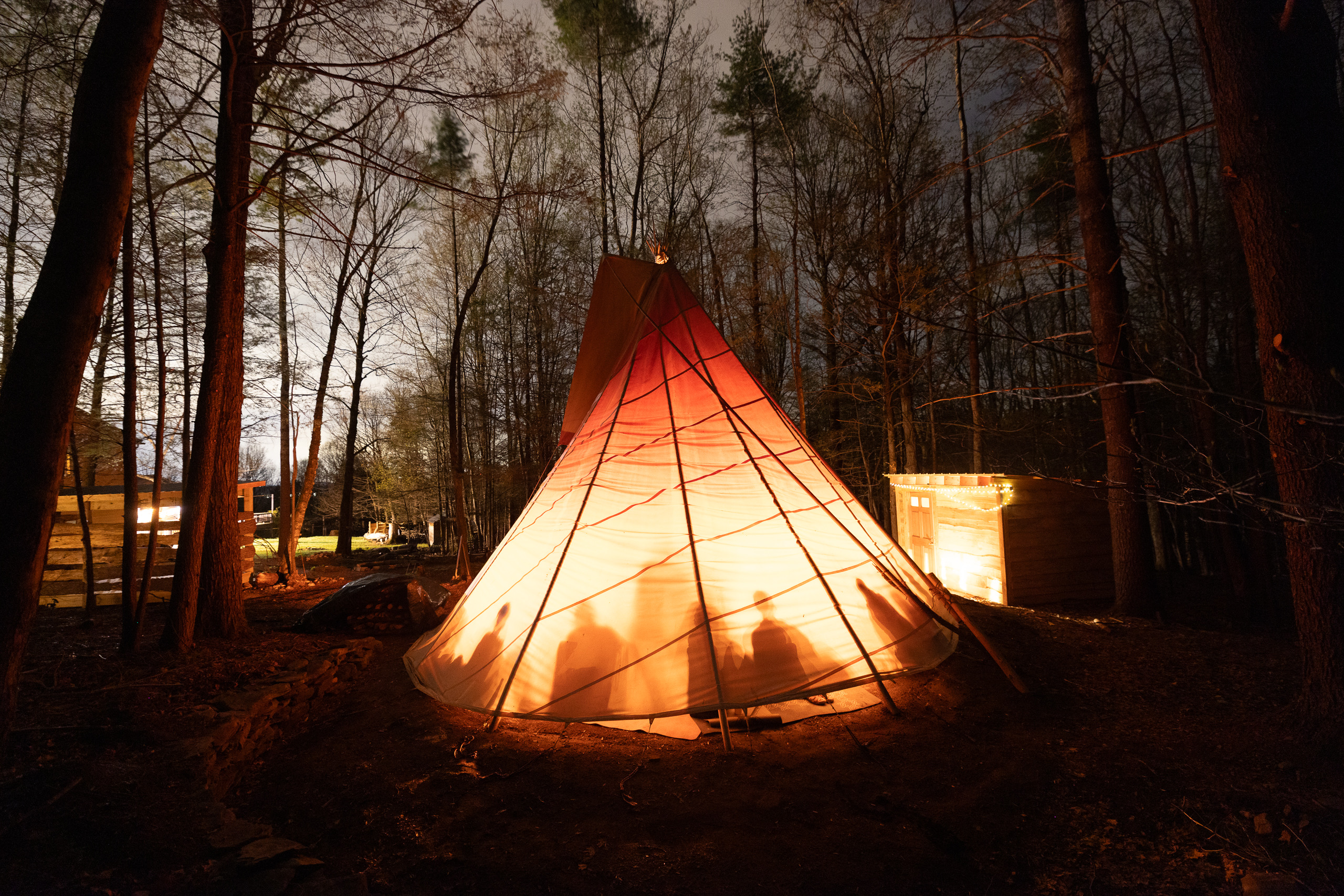 A tipi glowing at night in the woods, silhouettes inside